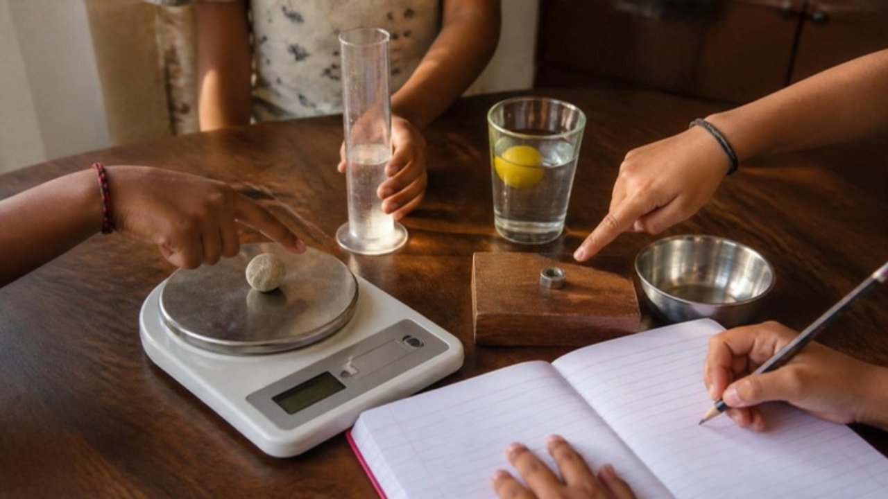 Padhai ki mez par digital taraazu jis par chhota pathar rakha hai, paas mein paani wala measuring cylinder, nimbu tairta hua paani ka glass, steel ki katori aur copy-pencil; bachche haath se ishaara kar rahe hain