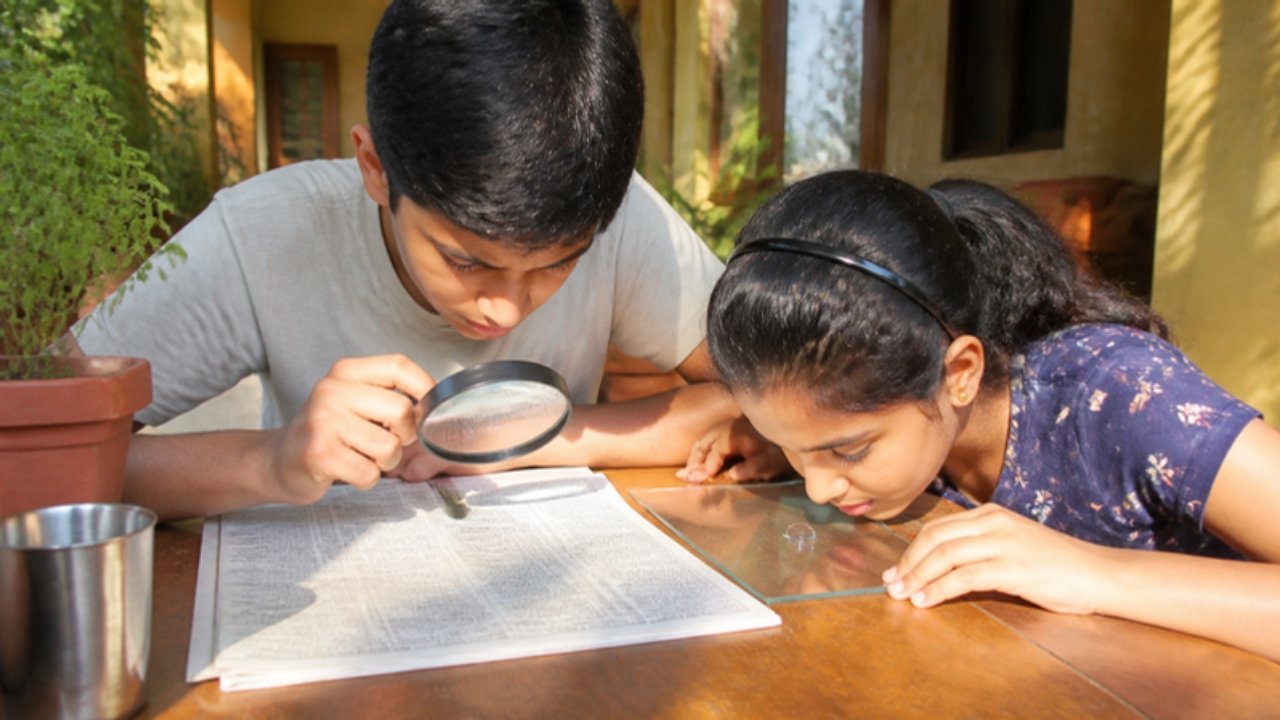 Verandah mein ek ladka magnifying glass se akhbaar ke akshar bade dekh raha hai aur ladki kaanch par rakhi paani ki boond se jhaank rahi hai