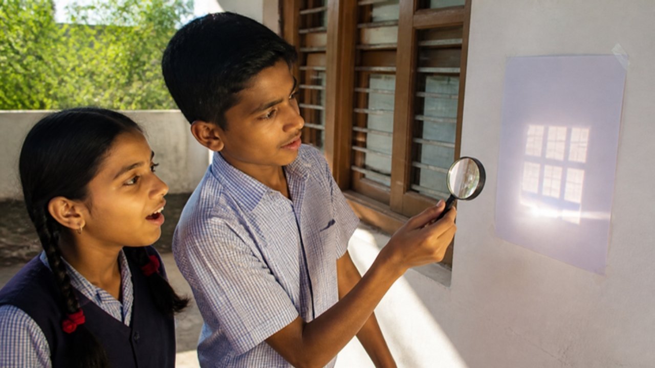 Verandah mein ek ladka magnifying glass pakde hai aur deewar par lage safed kaagaz par khidki ki chhoti chamakdaar image ban rahi hai, paas khadi ladki hairaan hokar dekh rahi hai