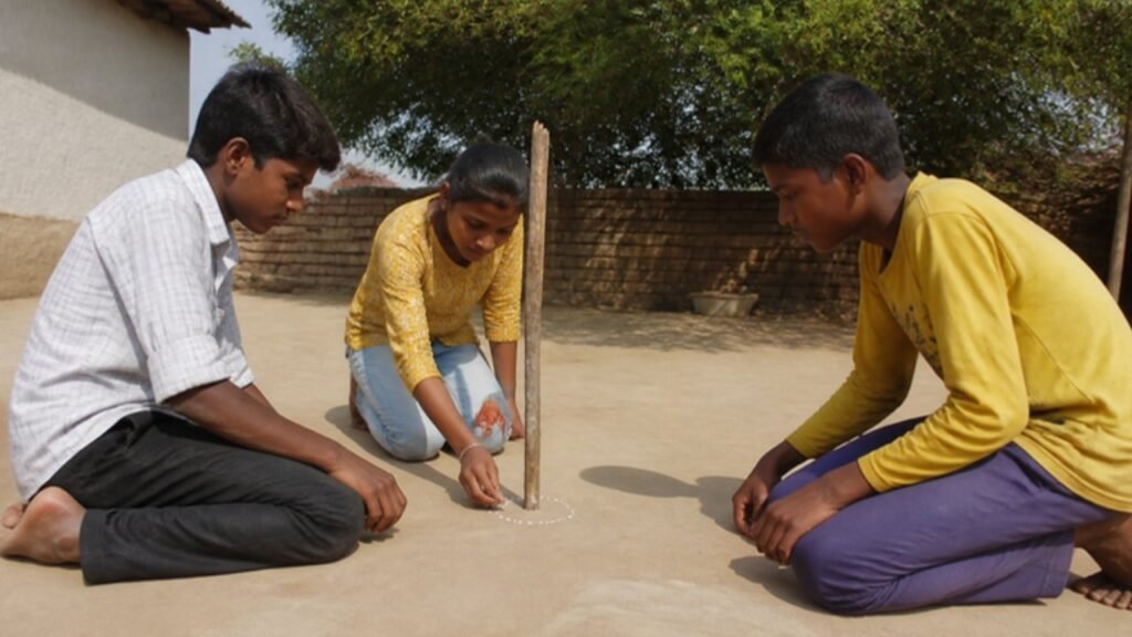Aangan mein teen bachche seedhi lakdi ki chhoti parchhai ki nok par chalk se bindu laga rahe hain