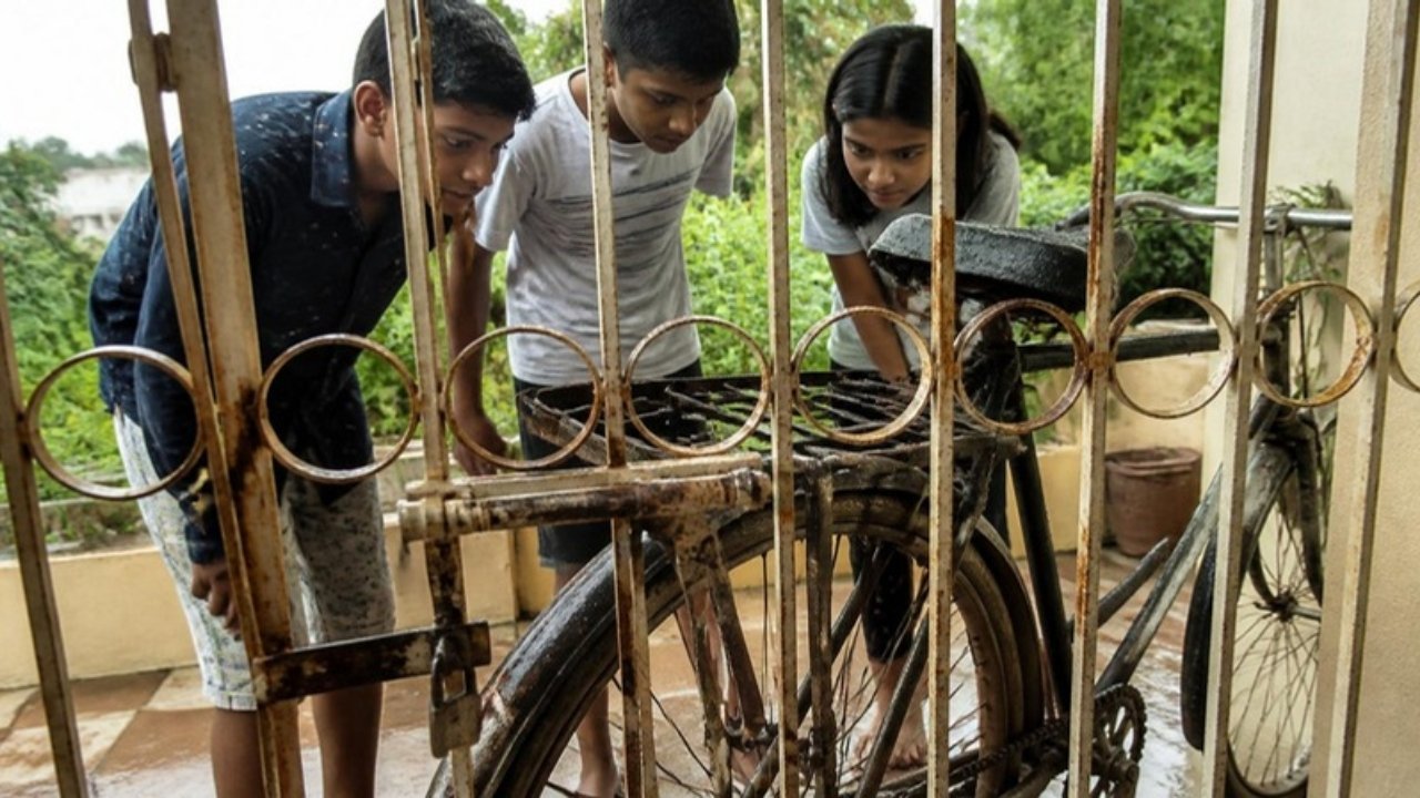 Barsaat ke mausam mein ghar ke jang lage gate ke peeche teen bachche jang lagi purani cycle ko dhyan se dekh rahe hain